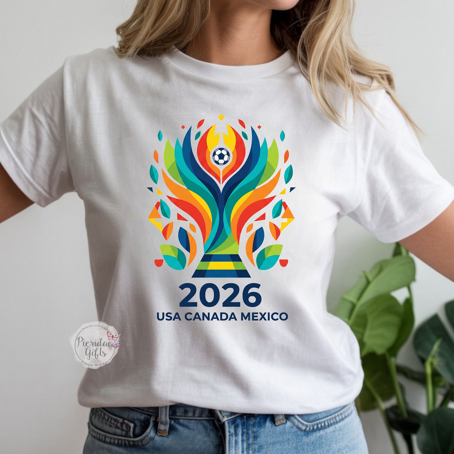 White t-shirt with colorful 2026 World Cup logo worn by a person, with a plant in the background.